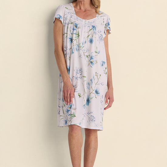 Woman wearing a lavender floral short nightgown with blue flowers, standing against a cream background.