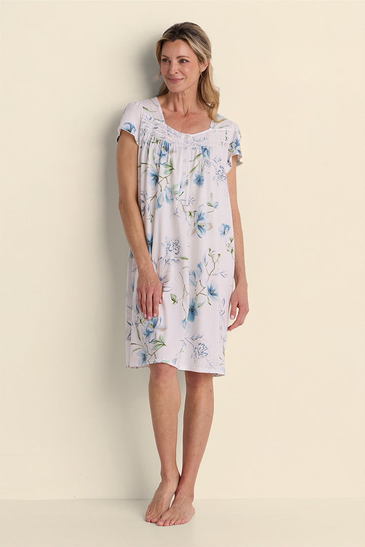 Woman wearing a lavender floral short nightgown with blue flowers, standing against a cream background.