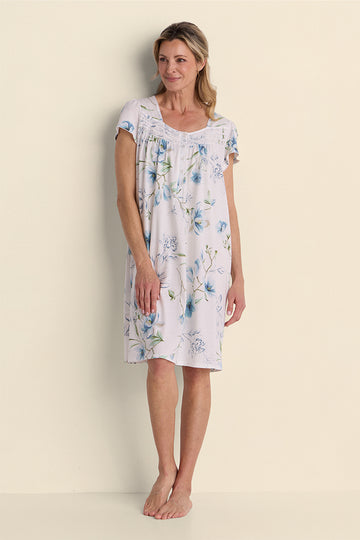 Woman wearing a lavender floral short nightgown with blue flowers, standing against a cream background.