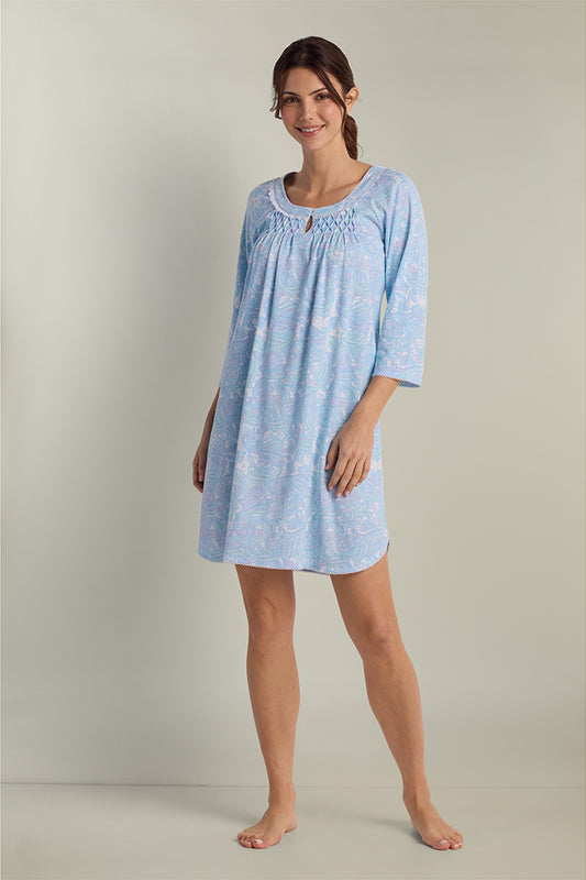 Woman wearing a short blue paisley nightgown with three-quarter sleeves and gathered neckline, standing against a neutral