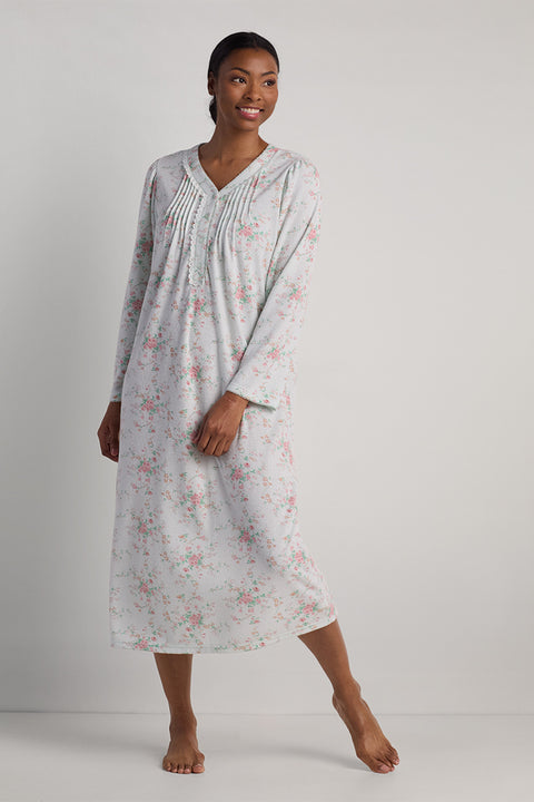 Woman wearing a long nightgown in light gray with pink floral pattern and pleated V-neckline, standing against neutral